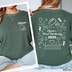 May include: Moss green t-shirt with white graphic. Front features "Mom" and a football helmet. Back has "Elijah's First Birthday 2025" with football-themed doodles. The shirt is a comfortable, casual style.