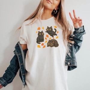 Może przedstawiać: Kremowy t-shirt z grafiką przedstawiającą trzy czarne koty z ż&oacute;łtymi oczami i jesiennymi liśćmi. Jeden kot siedzi, jeden jest w powietrzu, a jeden leży. Projekt jest wyśrodkowany na koszulce.