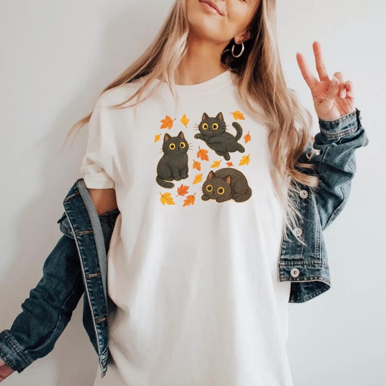 Może przedstawiać: Kremowy t-shirt z grafiką przedstawiającą trzy czarne koty z ż&oacute;łtymi oczami i jesiennymi liśćmi. Jeden kot siedzi, jeden jest w powietrzu, a jeden leży. Projekt jest wyśrodkowany na koszulce.