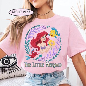 Puede incluir: Camiseta rosa claro con un gr&aacute;fico colorido de Ariel de La Sirenita, rodeada por una corona floral. El dise&ntilde;o incluye a Flounder, Sebasti&aacute;n y el texto "The Little Mermaid". La camiseta tambi&eacute;n tiene el texto "LIGHT PINK" en la parte superior izquierda.