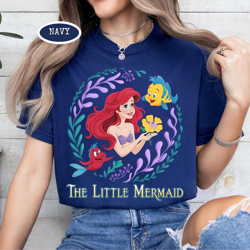 Puede incluir: Camiseta azul marino con una colorida ilustraci&oacute;n de Ariel de La Sirenita. El dise&ntilde;o incluye a Ariel, Flounder, Sebasti&aacute;n y una flor amarilla, rodeados por una corona de hojas. Las palabras "The Little Mermaid" est&aacute;n impresas debajo.