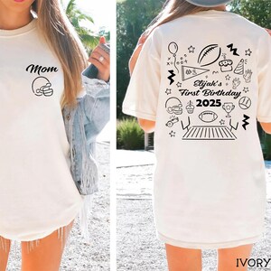 May include: Ivory-colored t-shirt with black graphic designs. The front features the word "Mom" and a football helmet. The back displays "Elijah's First Birthday 2025" with football-themed illustrations, including a football, cake, and stars.