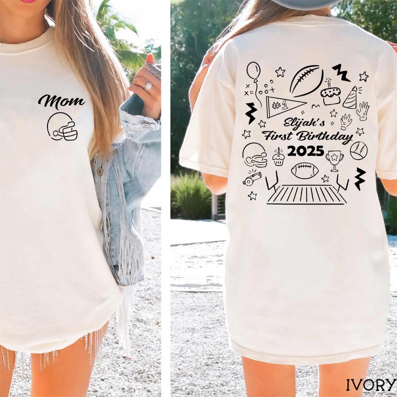 May include: Ivory-colored t-shirt with black graphic designs. The front features the word "Mom" and a football helmet. The back displays "Elijah's First Birthday 2025" with football-themed illustrations, including a football, cake, and stars.