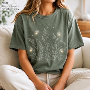 May include: A sage green short-sleeve t-shirt featuring a white line art graphic of tall grasses, wildflowers, and small flying insects. The design includes small white dots resembling light. Text reads: NOTE: This is a flat print, not actually glow.