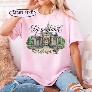 May include: Light pink t-shirt featuring a Disneyland castle illustration with ducks. The word "Disneyland" is written in cursive script above the castle. The shirt is a light pink color.