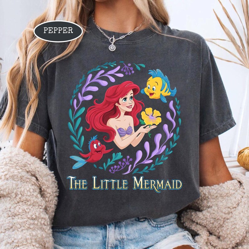 Puede incluir: Camiseta gris oscuro con una colorida ilustraci&oacute;n de Ariel de La Sirenita. El dise&ntilde;o incluye a Ariel con cabello rojo, un top de concha p&uacute;rpura y una flor amarilla, rodeada de personajes acu&aacute;ticos y el texto "The Little Mermaid."