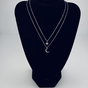 Può includere: Una collana d'argento esposta su un espositore nero. La collana presenta due fili, uno con un ciondolo a stella e l'altro con un ciondolo a mezzaluna. L'espositore è su una superficie bianca.