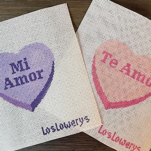 May include: Two needlepoint canvases with heart shapes. One heart is purple with "Mi Amor" text, the other is pink with "Te Amo" text. Both canvases have the text "Loslowerys" at the bottom.