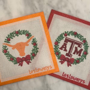 May include: Two needlepoint canvases with collegiate themes. One is orange with a Longhorn silhouette in a wreath, the other is red with "A&M" in a wreath. Both have a red bow and the text "LosLowerys".