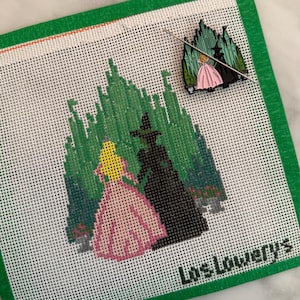 May include: Needlepoint canvas featuring a green forest scene with a princess in a pink dress and a black silhouette. A pin with a similar design rests on the canvas. The text "Los Lowerys" is at the bottom.