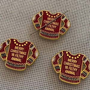 May include: Three enamel pins in the shape of a Christmas sweater. The pins are burgundy with gold trim and feature the text "MERRY CHRISTMAS YA FILTHY ANIMAL". A silver pin is resting on one of the pins.