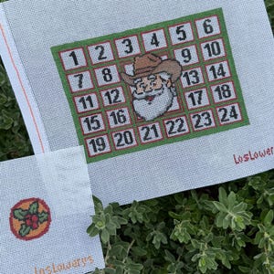 May include: Needlepoint canvas with a Christmas advent calendar design. The calendar features a grid of numbered squares, a Santa Claus wearing a cowboy hat, and the text "Los Lower". A smaller canvas shows a holly design.
