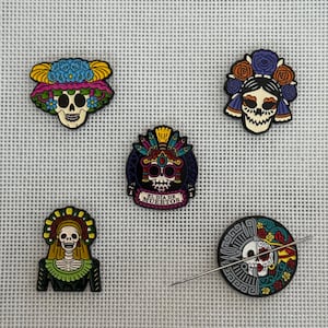 May include: A collection of Day of the Dead themed pins and a needle minder. The pins feature colorful sugar skull designs with floral and decorative elements. One pin has the text "EL DIA DE MUERTOS". The needle minder has a skull design.