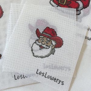 May include: White canvas with a cross-stitch design of Santa Claus wearing a red cowboy hat. The text "LosLowerys" is printed in black at the bottom. The image is a close-up of multiple canvases.