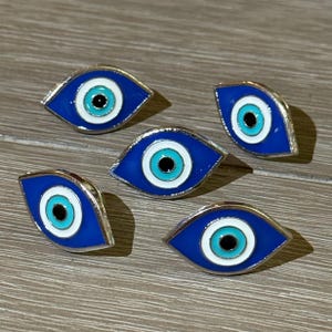 May include: Five blue enamel eye-shaped pins with a silver-colored border. Each pin features a white, light blue, and black center, resembling an eye. The pins are arranged on a light brown wooden surface.