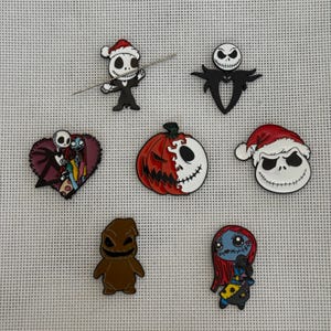 Puede incluir: Una colección de coloridos pines de esmalte con personajes de Pesadilla antes de Navidad. Incluye a Jack Skellington con gorro de Papá Noel, una calabaza, Sally y Oogie Boogie. Los pines están sobre un fondo gris claro texturizado.