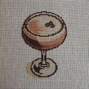 May include: Needlepoint design of a cocktail in a stemmed glass. The drink is a light brown color with a dark brown outline. The glass has a circular base and a detailed stem. The background is a white grid.