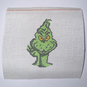 May include: A cross-stitch design featuring the Grinch, in shades of green with yellow eyes, on a white canvas. The Grinch is depicted with a mischievous expression, set against a neutral background. The canvas has a red stitched border.