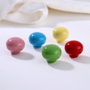 Könnte beinhalten: Fünf farbenfrohe Keramik-Schrankknöpfe in Rosa, Blau, Gelb, Grün und Rot. Jeder Knopf hat eine abgerundete Oberseite und einen kurzen, zylindrischen Sockel. Die Knöpfe sind auf einer weißen Oberfläche angeordnet, mit einem weichen, verschwommenen Hintergrund.