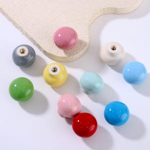 Op de afbeelding: Een verzameling kleurrijke keramische kastknoppen in verschillende vormen en maten. De knoppen zijn in tinten roze, blauw, groen, geel en rood. Sommige knoppen zijn rond, terwijl andere een meer unieke, afgeronde vorm hebben. De knoppen liggen op een wit oppervlak.