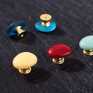 May include: A collection of colorful cabinet knobs with gold-colored bases. The knobs are in various colors, including blue, red, yellow, and green. The knobs have a smooth, rounded shape and are likely made of glass or resin.