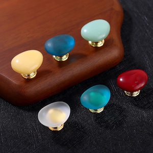 May include: A selection of colourful cabinet knobs with a smooth, oval design. The knobs come in various colours, including yellow, blue, green, and red, with a gold-coloured base. These decorative knobs are ideal for adding a pop of colour to furniture.