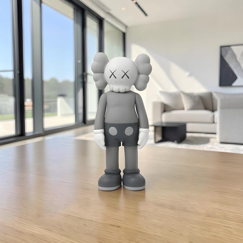 May include: A gray vinyl figure with a skull-like head featuring two black 'X' eyes and puffy ears. The figure wears gray shorts with two white circles and white gloves. The figure is standing on a wooden table in a modern living room.