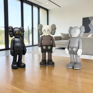May include: Three collectible vinyl figures in black, brown, and gray, each with distinctive X-shaped eyes and a rounded head. The figures stand on a wooden surface, showcasing a modern art aesthetic. The figures have white gloves and shorts with two circles.