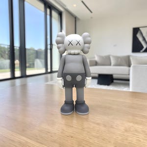 May include: A gray vinyl figure with a skull-like head and crossed-out eyes. The figure has gray shorts, a gray torso, and white gloves. The figure is standing on a wooden surface.