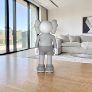 May include: A gray and white figurine with large, rounded ears and a blank face. The figure is wearing gray shorts and a matching shirt. The figurine is standing on a wooden surface in a well-lit room.