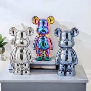 Puede incluir: Tres figuras decorativas de oso en plata, arcoíris iridiscente y azul metálico. Los osos tienen un detalle en forma de corazón en el pecho. La figura iridiscente destaca por sus colores vibrantes. Presentadas sobre una superficie gris.