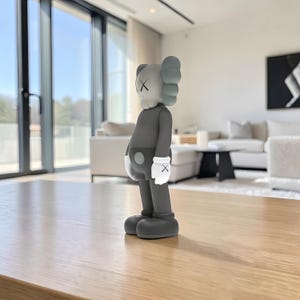 May include: A gray vinyl figure with a distinctive design. The figure has a gray body, a white head with black 'X' eyes, and light blue ears. It wears white gloves with black 'X' marks. The figure stands on a wooden table in a well-lit room.
