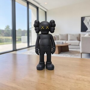 May include: A black vinyl figure with a distinctive design. The figure has a rounded head with two large, puffy ear-like shapes and yellow 'XX' eyes. It is wearing shorts and has rounded feet. The figure is standing on a wooden table.