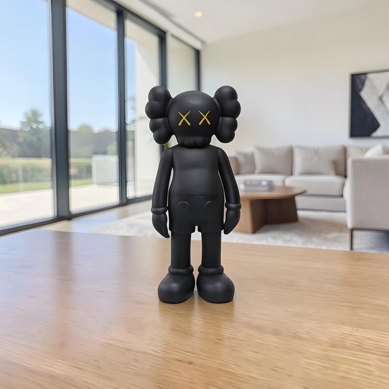 May include: A black vinyl figure with a distinctive design. The figure has a rounded head with two large, puffy ear-like shapes and yellow 'XX' eyes. It is wearing shorts and has rounded feet. The figure is standing on a wooden table.