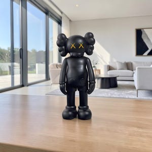 May include: A black vinyl figure with a rounded head, large ears, and crossed-out eyes. The figure has a smooth, matte finish and stands on a wooden surface. The figure is a collectible art toy.