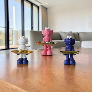 May include: Three colorful figurines, each holding a small, gold-toned tray. The figurines are in white, pink, and blue, with large, cartoonish eyes. The figurines are standing on a wooden table.