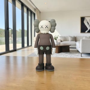 May include: A brown and gray vinyl figure with a skull-like face and crossed-out eyes. The figure has gray ears, white gloves, and black shorts with two green dots. It stands on a wooden surface.