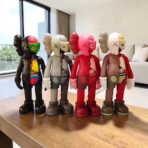 May include: Four KAWS-style vinyl figures in a row, each with a unique color scheme. The figures are split down the middle, revealing internal anatomy on one side. The color palette includes black, gray, pink, and brown, with contrasting internal colors.