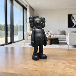 May include: A black vinyl figurine with yellow 'XX' eyes and rounded ears stands on a wooden surface. The figure has a stylized human form with large feet and hands. The background shows a modern living room.