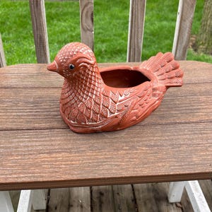 May include: A terracotta bird-shaped planter with detailed feather and wing patterns. The bird has a small opening for planting and is sitting on a wooden surface. The planter is a decorative item for gardens or indoor spaces.