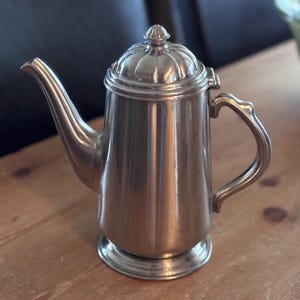 May include: A silver-colored metal coffee pot with a curved spout, a handle, and a lid with a small knob. The pot sits on a round base. The pot is on a wooden surface.