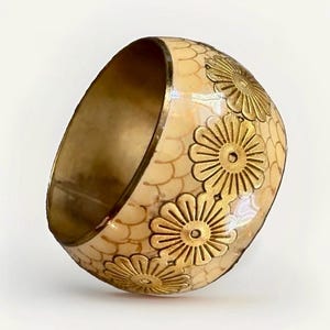 Könnte beinhalten: Ein breiter Armreif mit cremefarbener Oberfläche und einem feinen, sich wiederholenden Schuppenmuster. Die Außenseite ist mit einer vertikalen Reihe aus drei erhabenen, goldfarbenen Blumenmotiven verziert. Die Innenseite besteht aus glattem Messing.