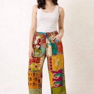 May include: Multi-coloured patchwork trousers featuring a variety of patterns in shades of red, yellow, green, and orange. The trousers have a drawstring waist and a relaxed fit. The model is wearing a white vest top.