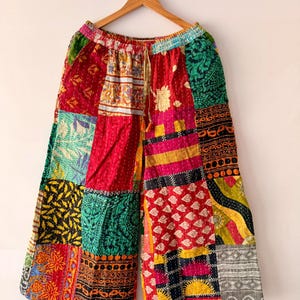 May include: Patchwork trousers in a vibrant mix of colours and patterns, including red, green, and yellow. The trousers have a drawstring waist and are displayed on a wooden hanger. The background is white.