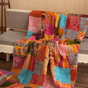 May include: A vibrant patchwork quilt in shades of pink, blue, yellow, and orange, draped over a grey sofa. The quilt is made from many fabric squares sewn together. A decorative pillow is on the left. The quilt is a mix of patterns.