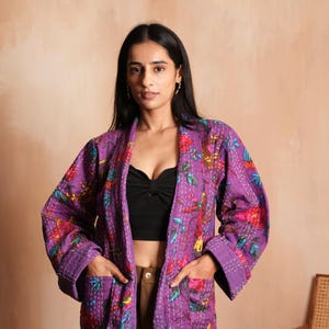May include: A purple quilted jacket with a floral pattern in red, pink, and green. The jacket has pockets and white stitching. The model is wearing a black top and brown pants.