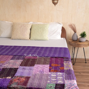 May include: A patchwork quilt in shades of purple, with a variety of patterns and textures. The quilt is on a bed with white pillows and a wooden headboard. The text "Kantha Way" is visible on the quilt.