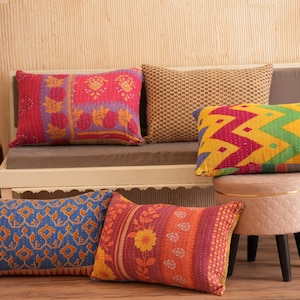 Op de afbeelding: Een verzameling decoratieve sierkussens in diverse kleuren en patronen. Kussens hebben bloemen-, geometrische en zigzagontwerpen. De kussens zijn gerangschikt op een bank en een kleine kruk, met een houten vloer en een houten lamp op de achtergrond.