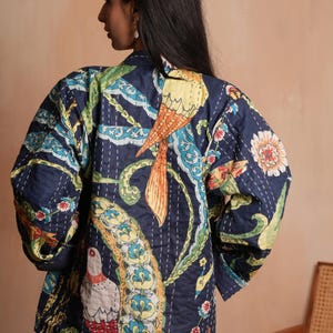 May include: A navy blue quilted jacket with a colourful bird and floral pattern. The jacket features a design with yellow, orange, green, and blue accents. The stitching adds texture to the garment.