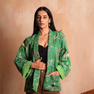 May include: Green kimono jacket with a jungle print featuring birds and tigers. The jacket has a contrasting green trim and two front pockets. The model is wearing a black top and brown pants.